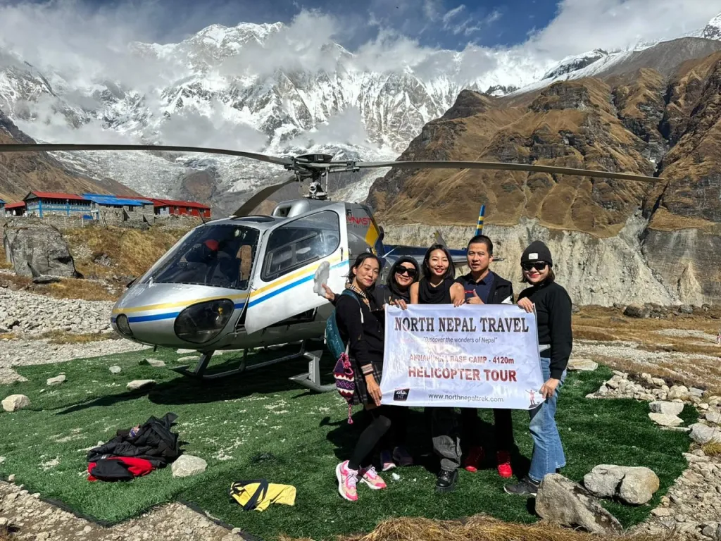 Five Clients, 1 male 4 female, from North Nepal trek clicking photos with banner of company helicopter just behind, with mountains in the background during ABC helicopter tour